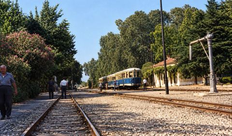 Colour print (colours restored) of the platform and tracks at Myloi station with a railcar under the trees.