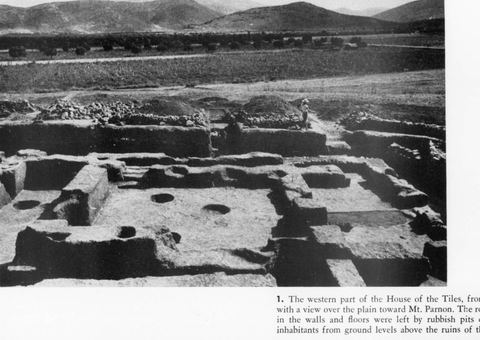 The western part of the House of the Tiles at Lerna from the north, with a view over the plain with olive groves, a dirt road and the mountains, and an excavation worker on the edge.