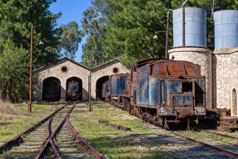 'Metre gauge graveyard. Mili Nafpliou Depot': the engine shed with arched doors at left, two water towers (stone base, blue tank) at right and tender locomotive 7723 in front.