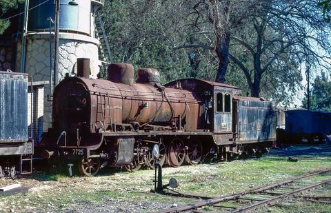 'Class Es no.7725 dumped at Mili Nafpliou depot': locomotive 7725 with a water tower behind and part of a stone building at left.