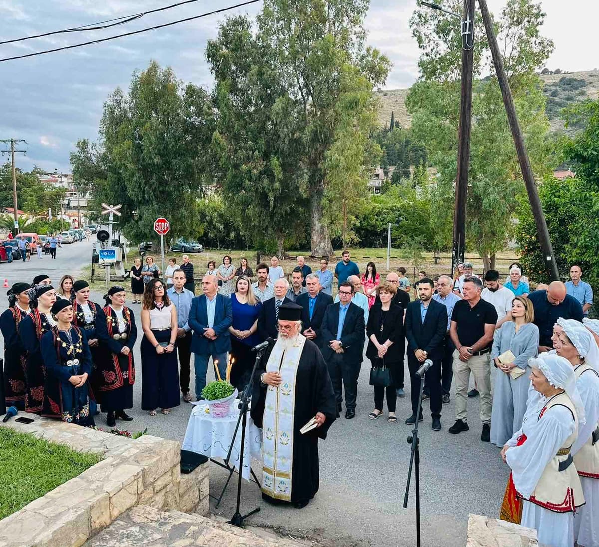 Villagers, guests and women in traditional costume stand around the priest during the memorial service by the old railway line, 15 June 2025