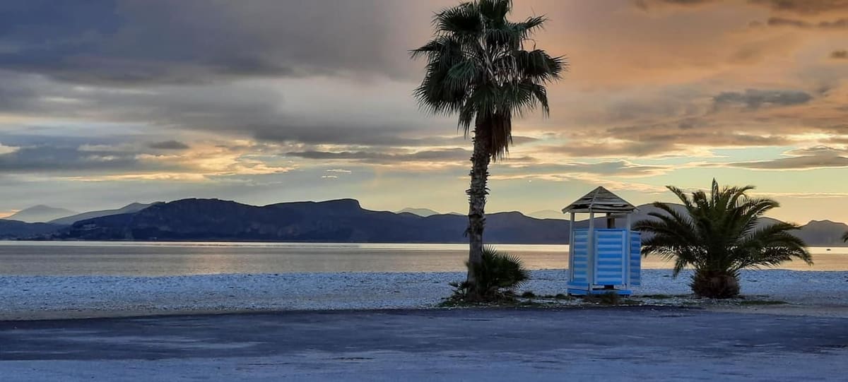 Myloi beach at dusk with palm trees and a lifeguard hut, mountains across the gulf