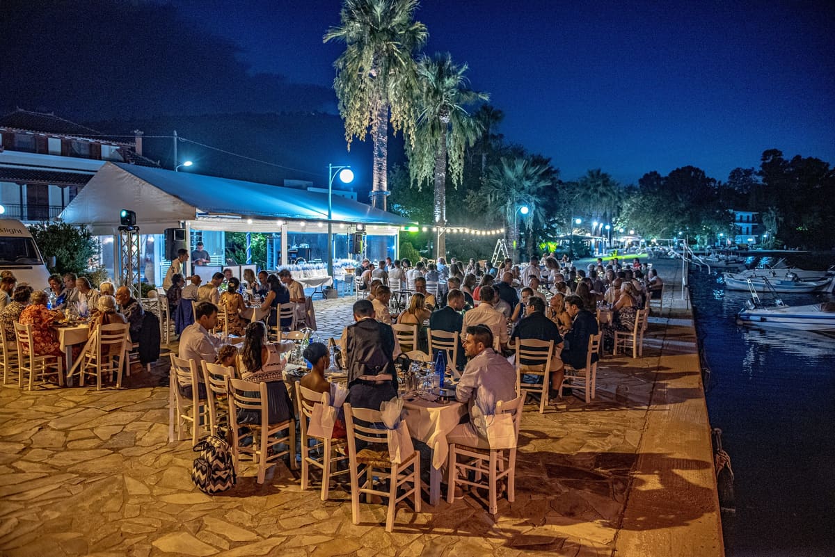 Long dinner tables full of guests on the stone quay at Faros at night, under the palm trees by the harbour