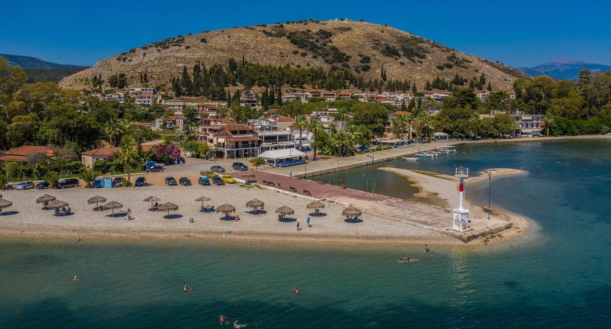 Luchtfoto van Myloi: kiezelstrand, pier met vuurtoren en de boulevard, met achter het dorp de heuvel Pontinos en de kasteelruïne