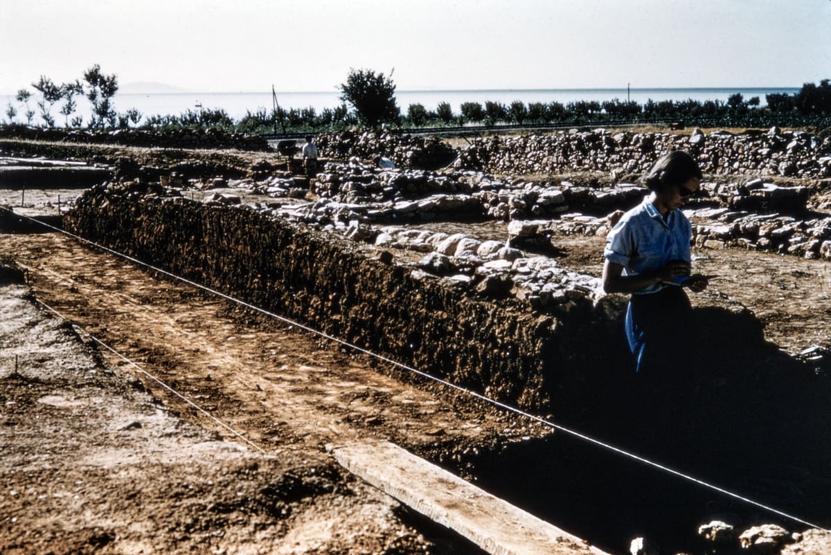 A member of the 1950s excavation team stands in a trench at Lerna, writing notes, with a measuring string, stone walls and the gulf behind