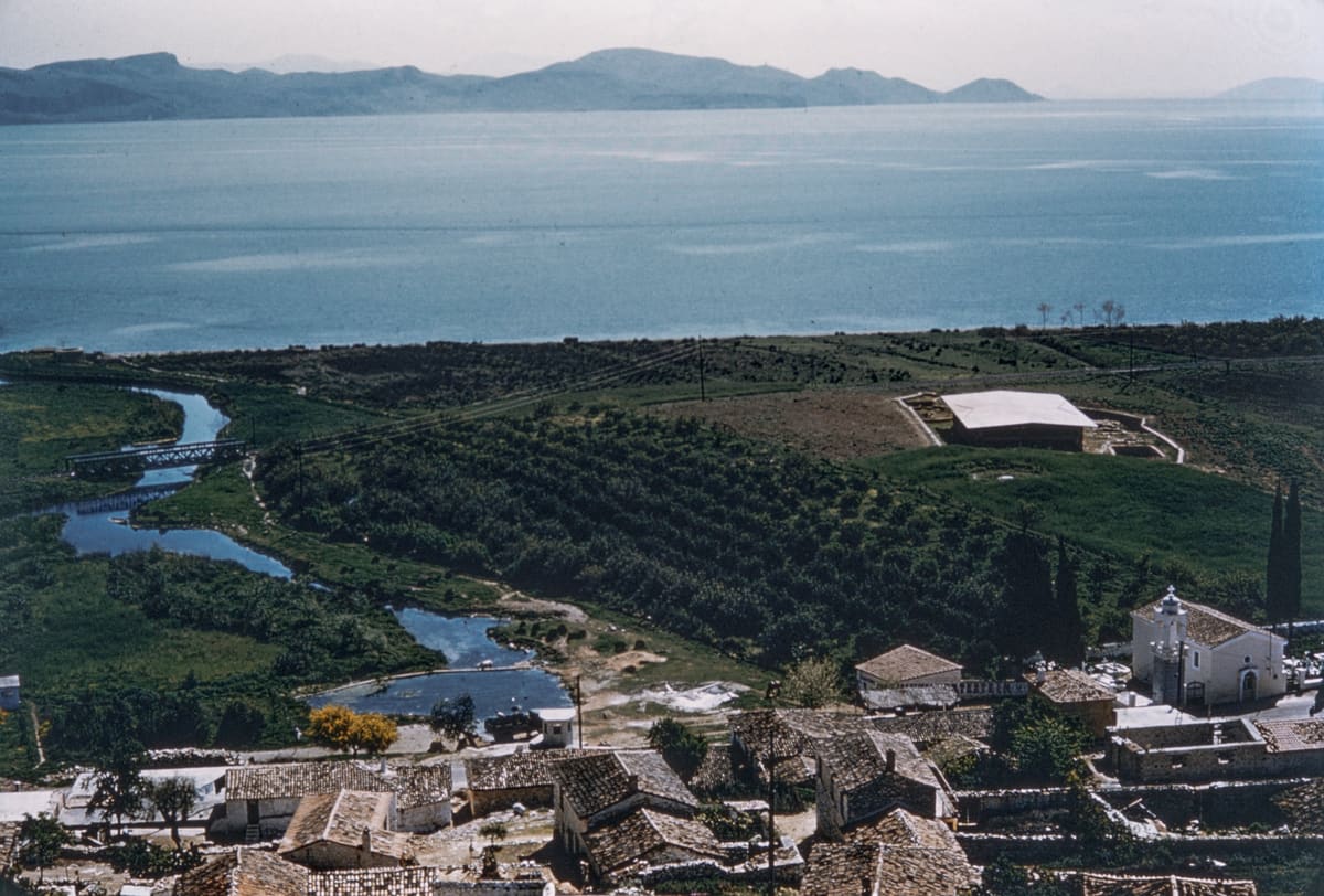 View over Myloi in the 1950s: tiled roofs and a church with a bell tower in front, the winding stream from the Lerna springs, the new roof over the excavation among the fields, and the gulf beyond