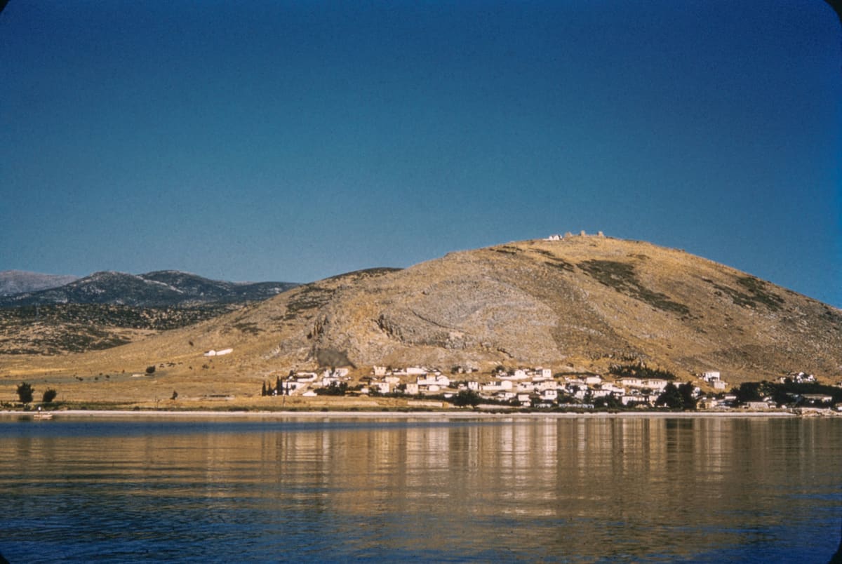 Οι Μύλοι τη δεκαετία του 1950 από τη θάλασσα: λευκά σπίτια στην ακτή στους πρόποδες του γυμνού λόφου του Ποντίνου, με τα ερείπια του κάστρου στην κορυφή, καθρεφτίζονται στα ήρεμα νερά του κόλπου