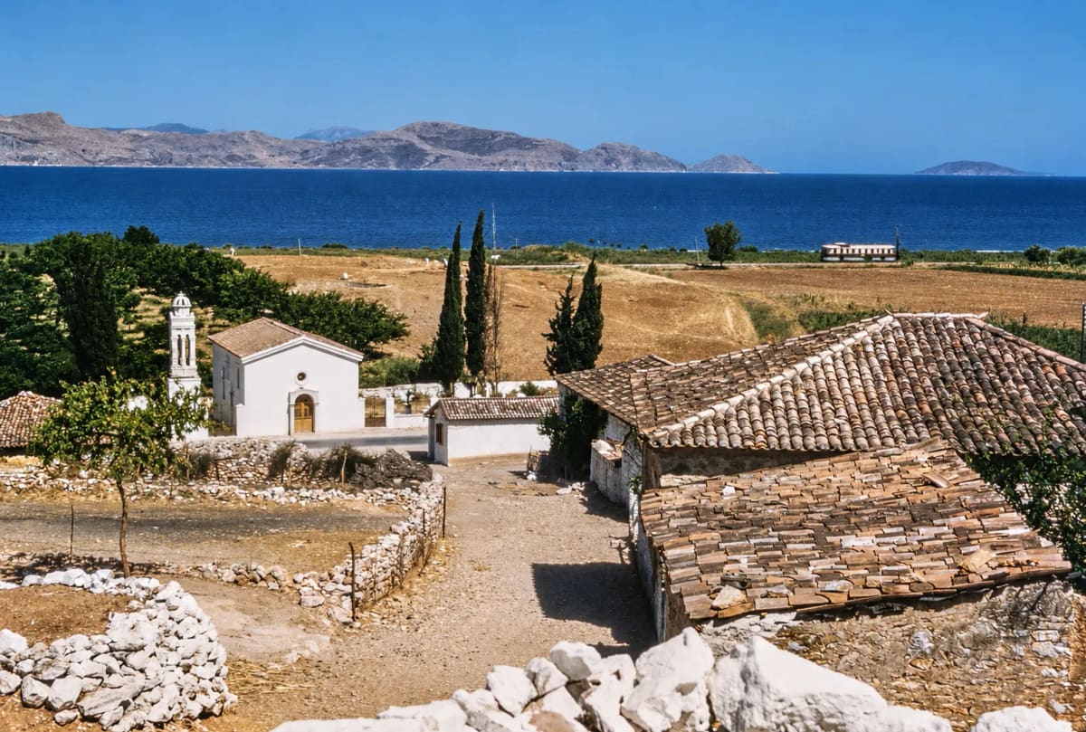 The church of Agios Dimitrios with its bell tower and cypresses at the edge of Myloi in the 1950s, with tiled roofs in front and the fields, a railcar and the gulf beyond