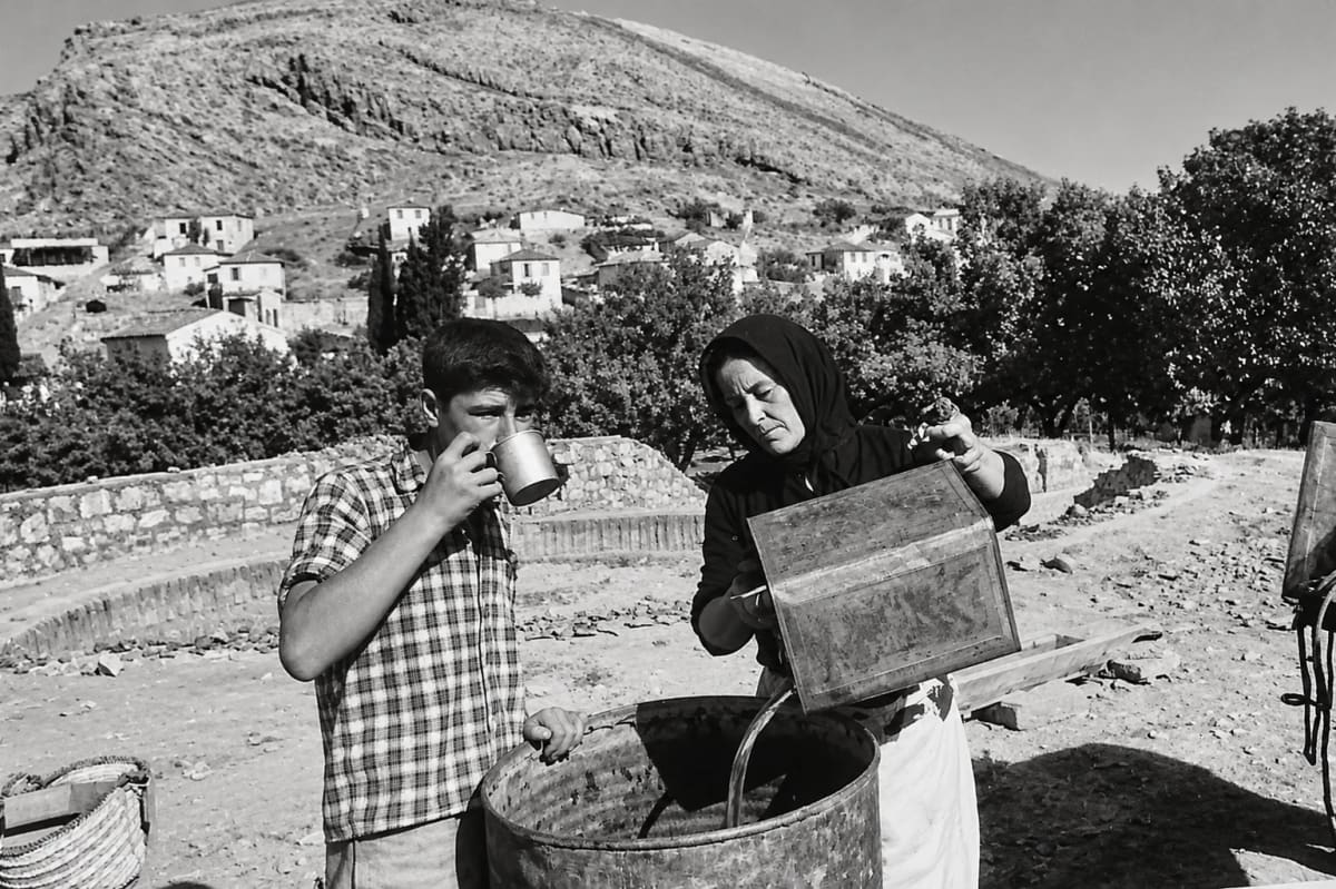 Members of the 1950s Lerna excavation team: a boy drinking from a tin mug and a woman in a headscarf pouring water into a barrel, with the houses of Myloi and Pontinos behind (digitally restored)