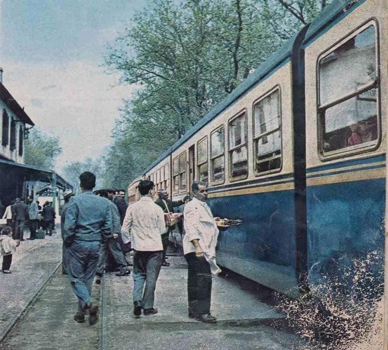 Obers in witte jasjes dragen dienbladen met souvlaki langs een blauwe trein op station Myloi