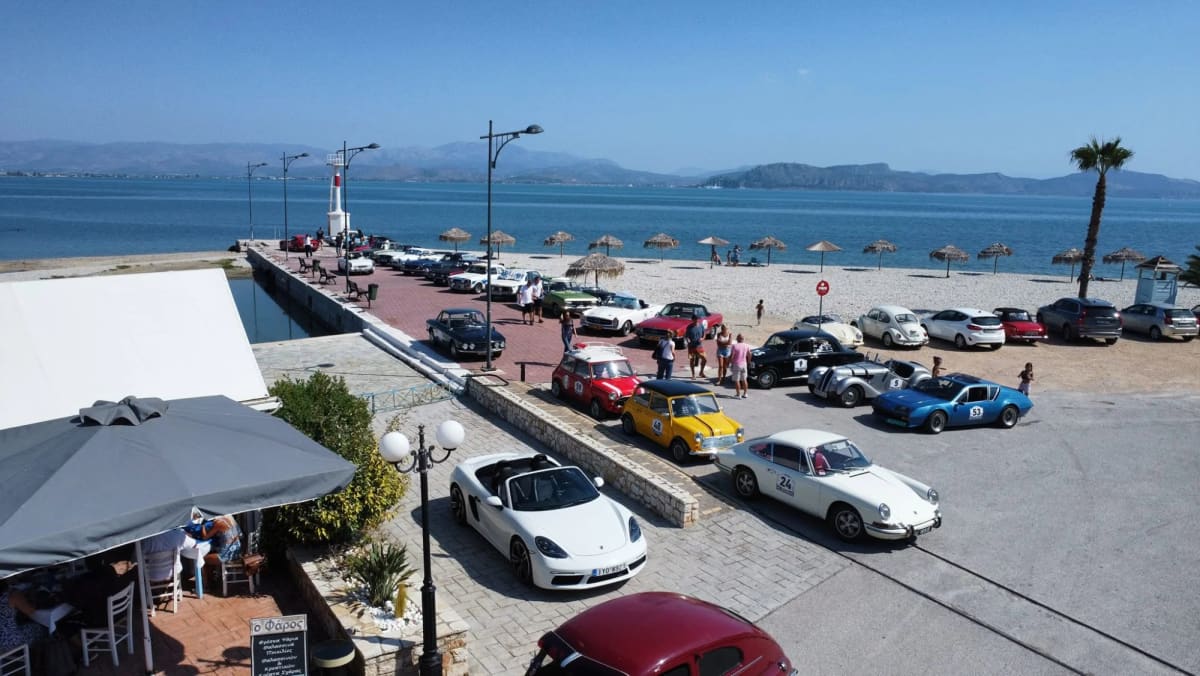 Κλασικά αυτοκίνητα ράλι με αριθμούς παρκαρισμένα στον μόλο και στην παραλιακή των Μύλων, από ψηλά, με τον φάρο και τον κόλπο πίσω