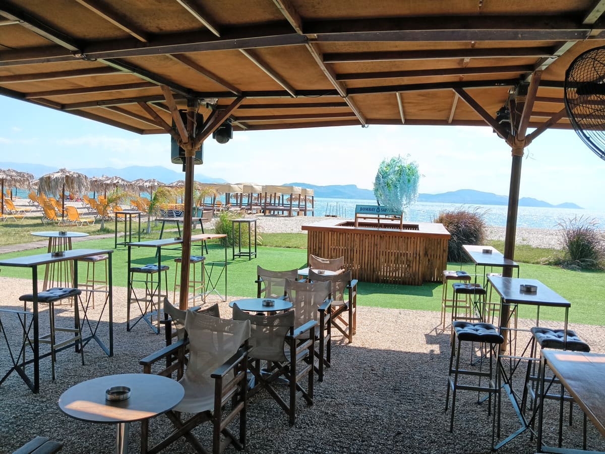 Shaded bar terrace with high tables and director's chairs, a wooden bar on the lawn and umbrellas on the beach beyond