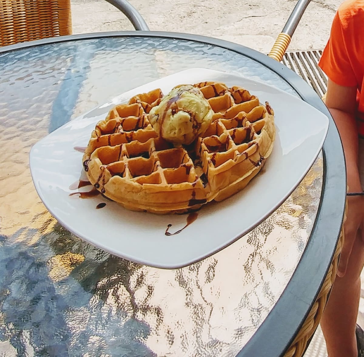 A waffle with a scoop of ice cream and chocolate sauce on a glass terrace table