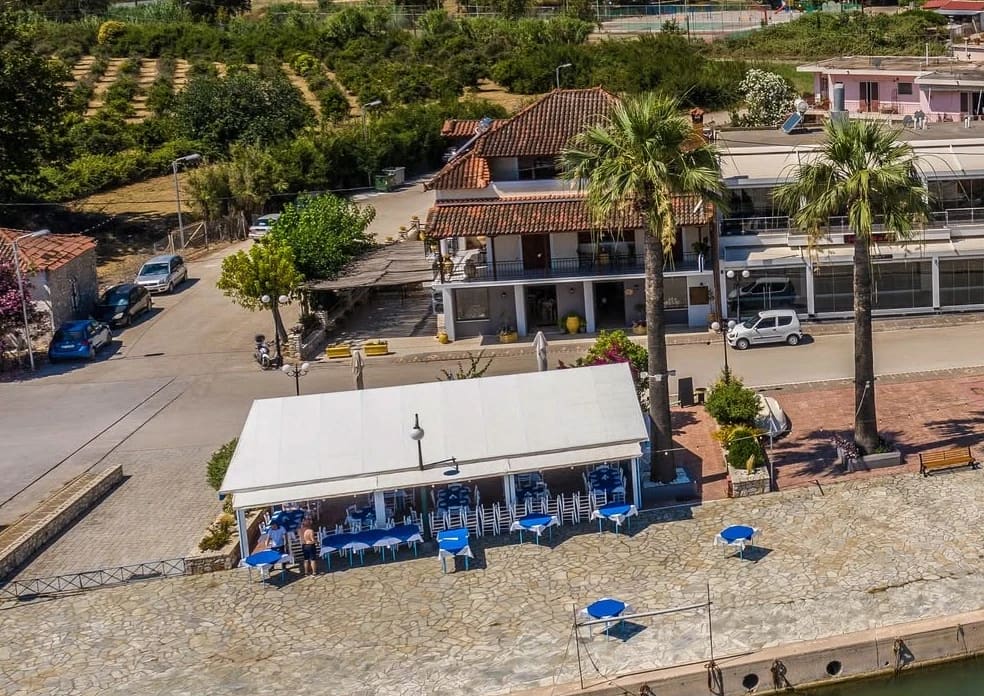 Faros vanuit de lucht: het overdekte terras en de blauwe tafels op de geplaveide kade aan het water, het huis aan de overkant van de weg en twee hoge palmen