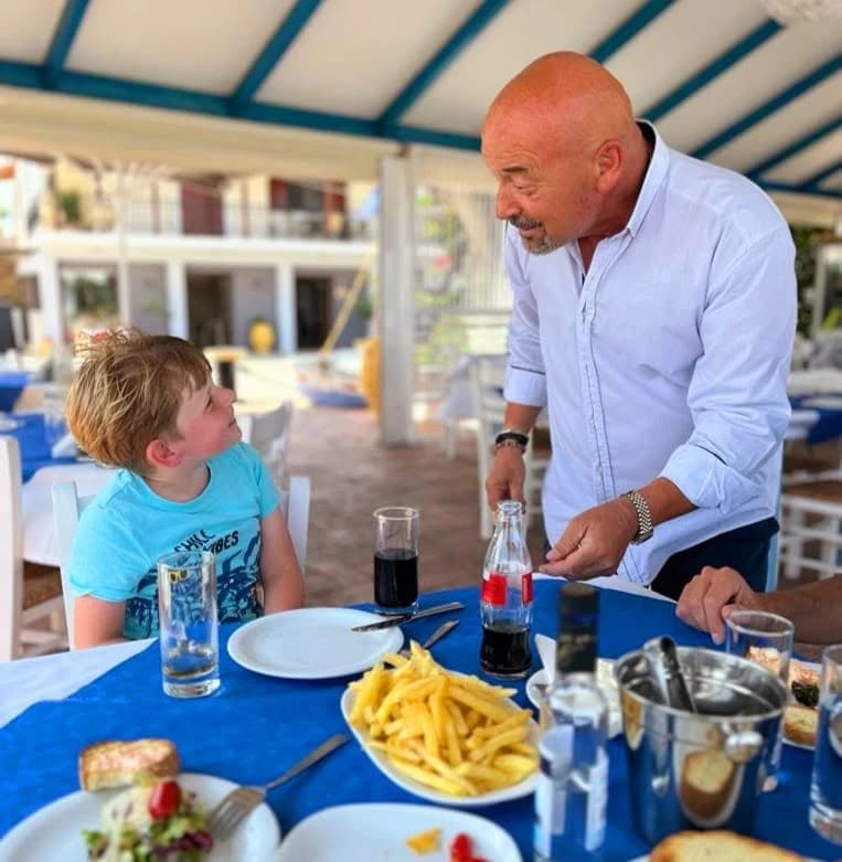 Kostas, eigenaar van Faros, in gesprek met een jonge gast aan een tafel onder het zonnescherm van de taverna