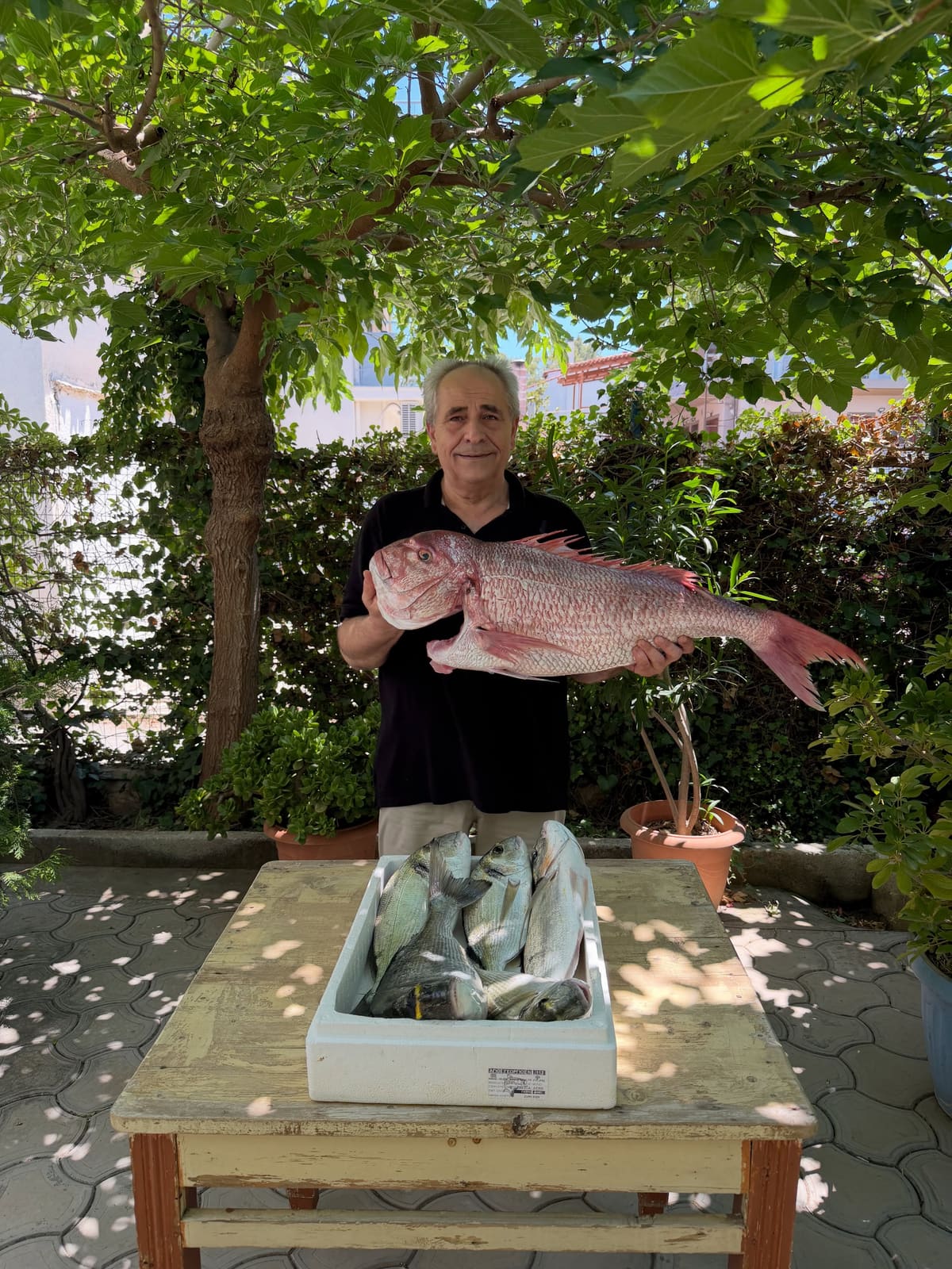 A man holds up a large red snapper behind a box of fresh fish under the trees outside Ta 5F