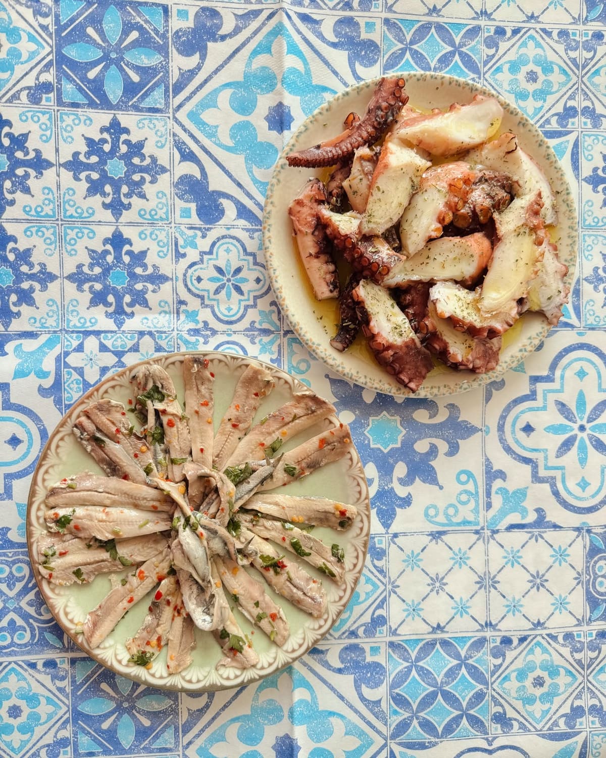 Two plates on a blue patterned table: grilled octopus and marinated white anchovies with herbs