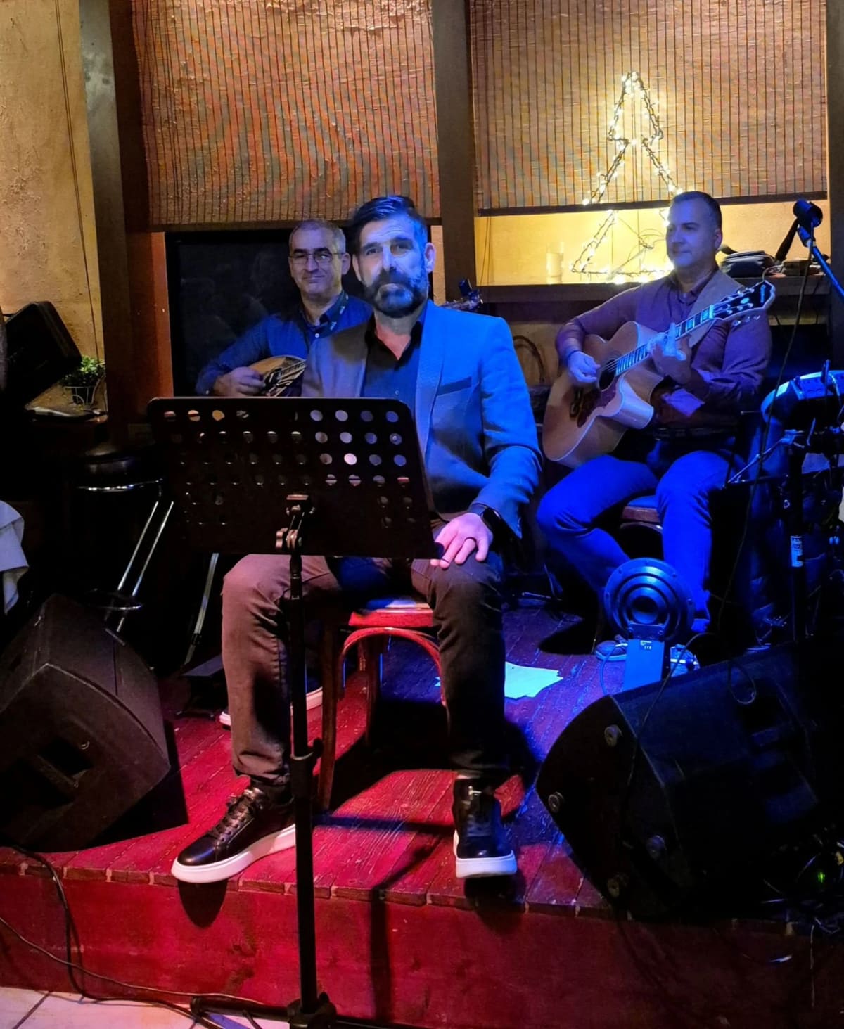 Drie muzikanten op het podiumpje van Grammes: een zanger achter een muziekstandaard, een bouzoukispeler en een gitarist