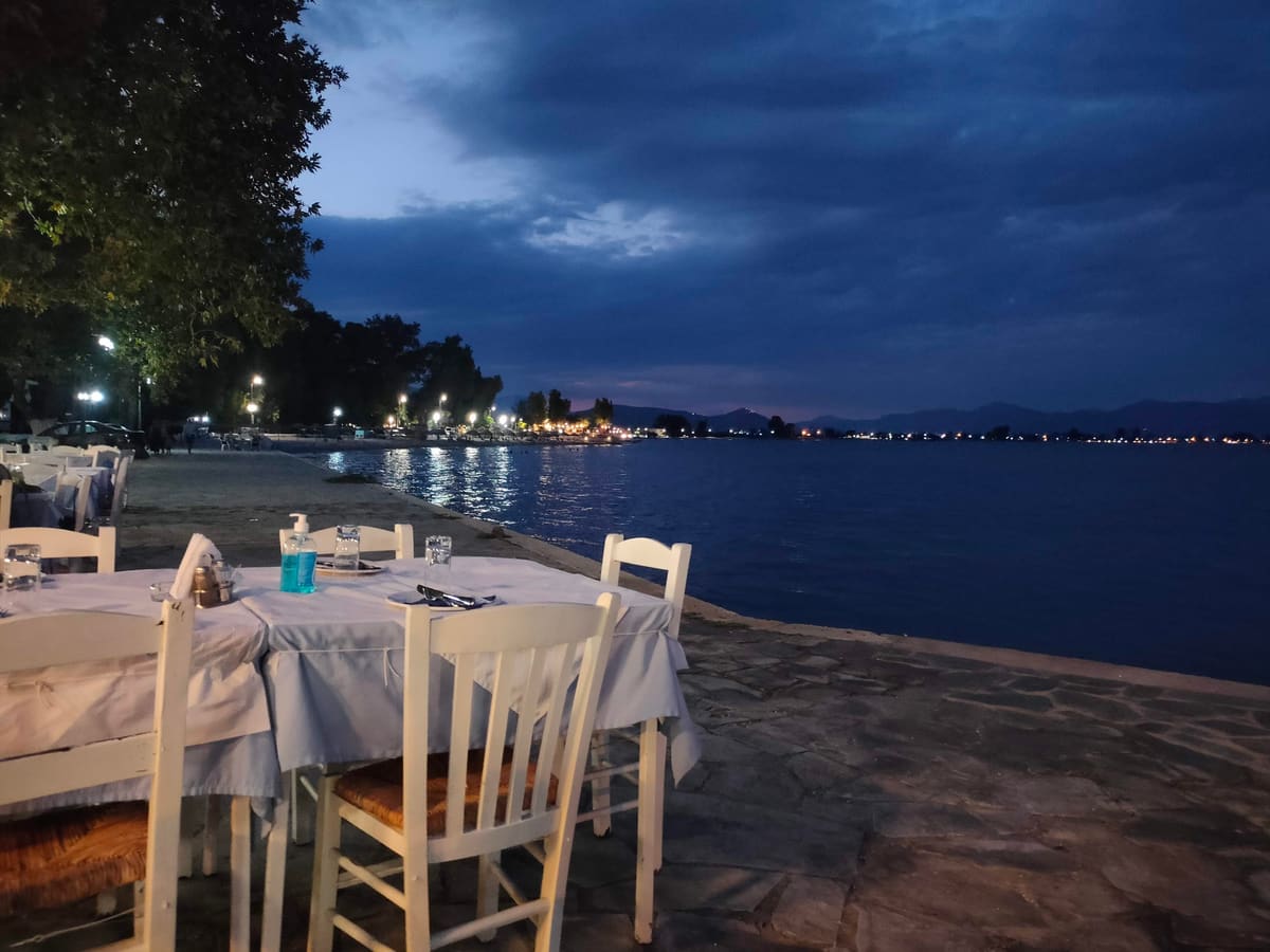 White taverna tables on the stone quay at dusk, beside the calm sea, with the lights of the Myloi waterfront reflected in the water