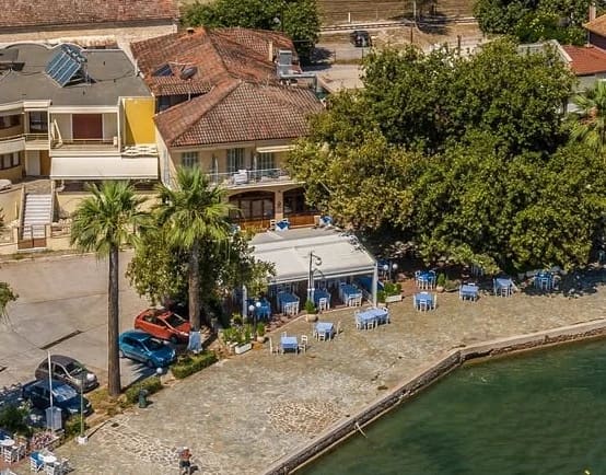 Ο Λύκος από ψηλά: η ταβέρνα με τη λευκή τέντα και τα μπλε τραπέζια στην πλακόστρωτη προκυμαία δίπλα στη θάλασσα, φοίνικες και ένα μεγάλο σκιερό δέντρο