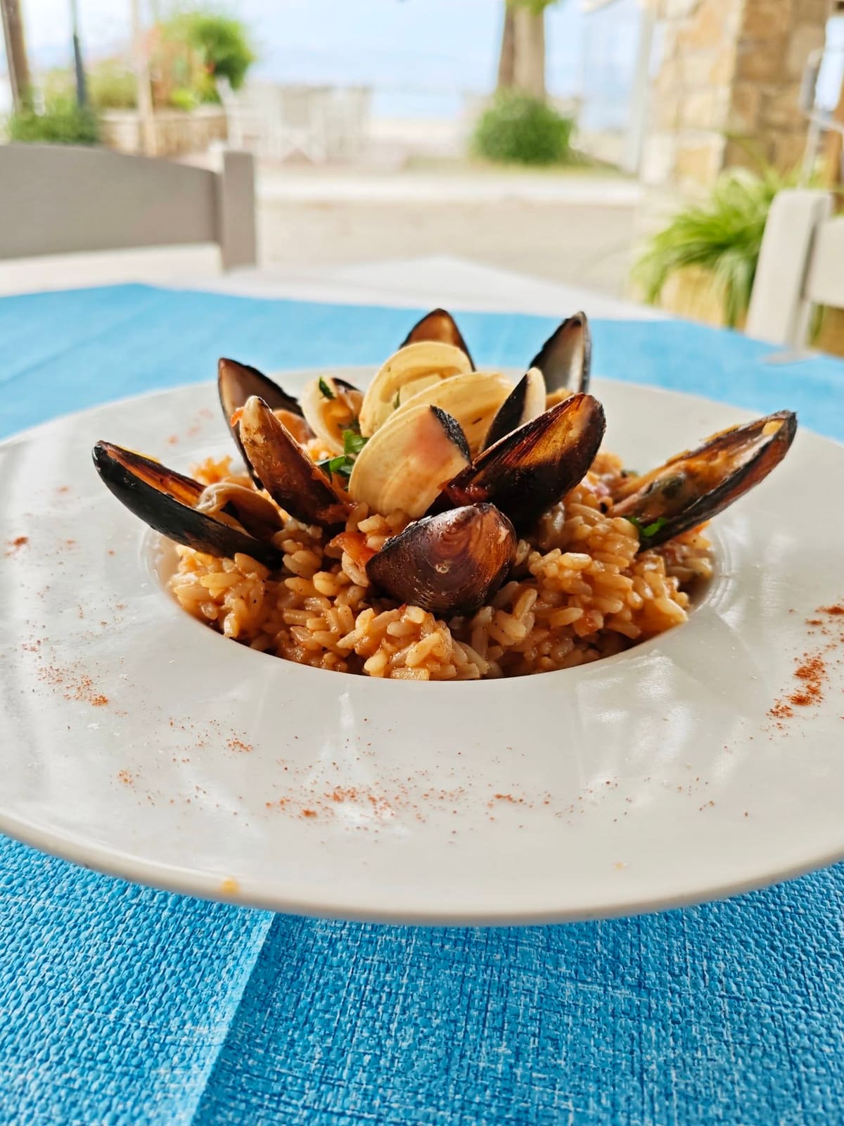 Talerz risotto z małżami na niebieskim obrusie, a za nim plaża i morze