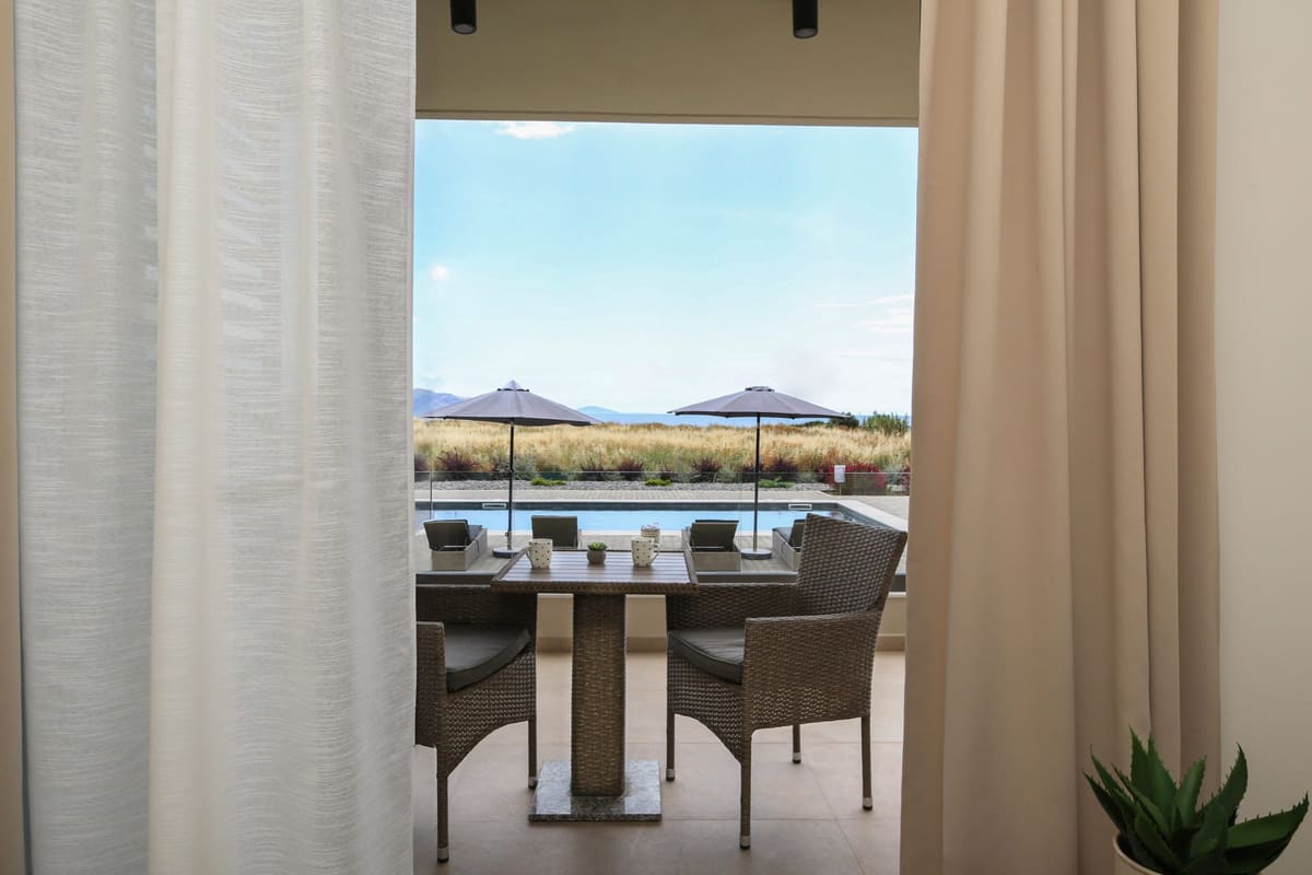 A terrace with a table and wicker chairs, seen through light curtains, with the garden beyond