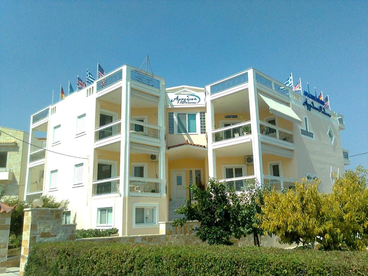The white and yellow three-storey Argoliki Thalassa building with its name sign, flags on the roof and balconies on every floor, behind a green hedge