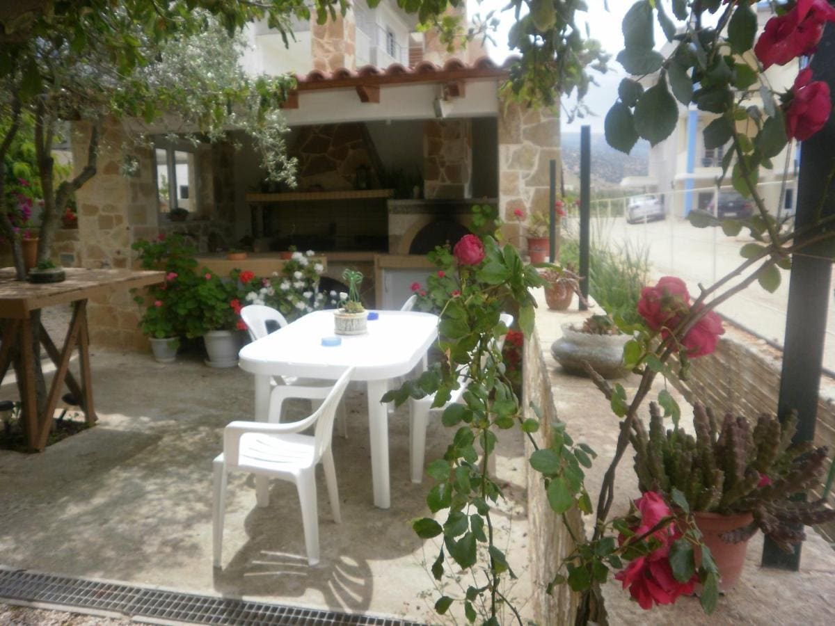 Σκιερή αυλή με λευκό τραπέζι και καρέκλες, τριαντάφυλλα και φυτά, και πίσω μια πέτρινη εξωτερική κουζίνα