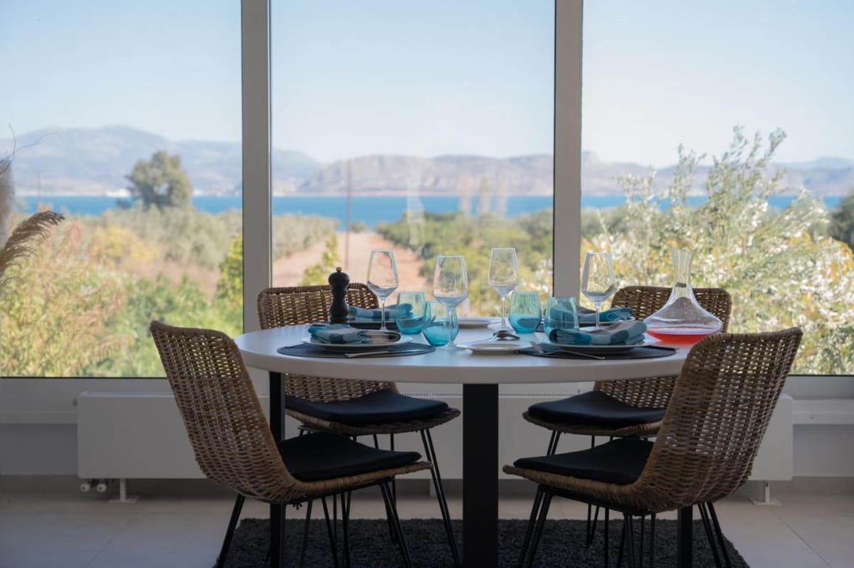 Round dining table set for four by a large window, with olive trees and the Argolic Gulf beyond