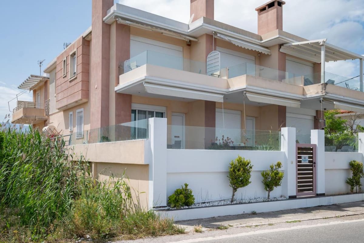 Το σπίτι από τον δρόμο: ροζ και λευκοί τοίχοι, γυάλινα μπαλκόνια με τέντες και λευκός μαντρότοιχος με μικρά δέντρα