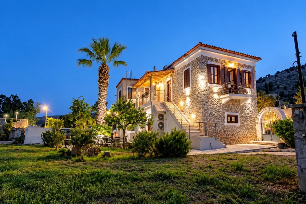 The stone house at dusk with warm wall lights, an outside staircase to the upper floor, a tall palm tree and a lawn in front