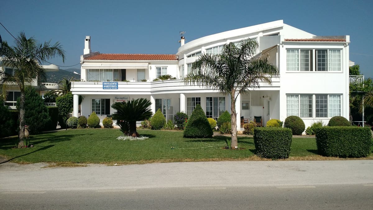 The white two-storey Miloi Rooms and Apartments building with a curved upper floor, a lawn with palm trees and clipped shrubs, and the reception sign