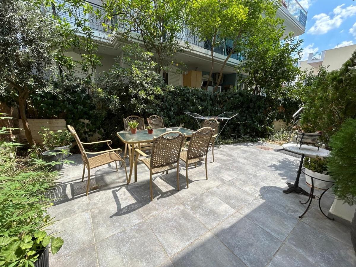 Paved garden courtyard with a table and wicker chairs among trees and plants, with the apartment building behind