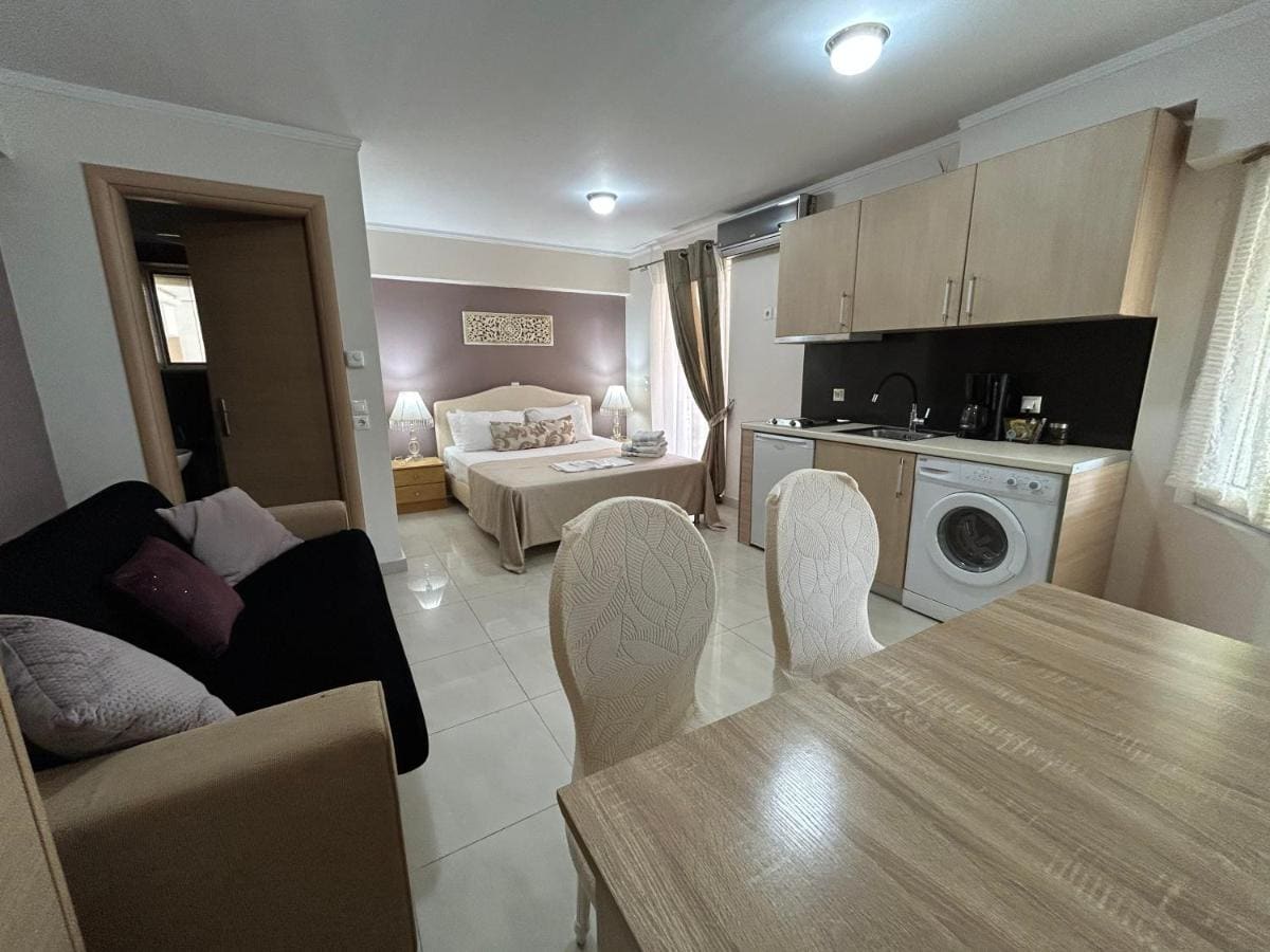 Open appartement met keuken en wasmachine, een eettafel met witte stoelen, een bank en verderop het bed
