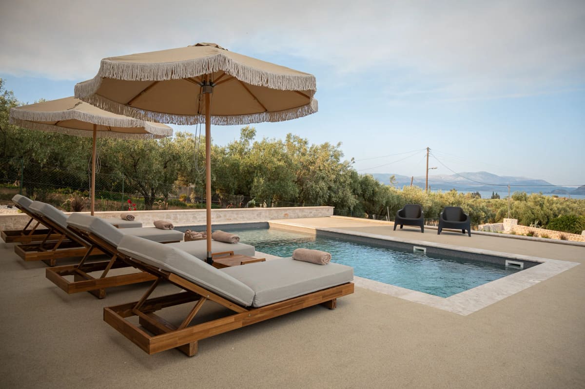 Sun loungers with fringed parasols beside the private pool, olive trees around and the gulf and mountains in the distance