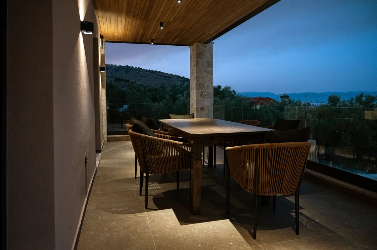 The covered terrace with a dining table and chairs at dusk, looking out over olive trees to the sea